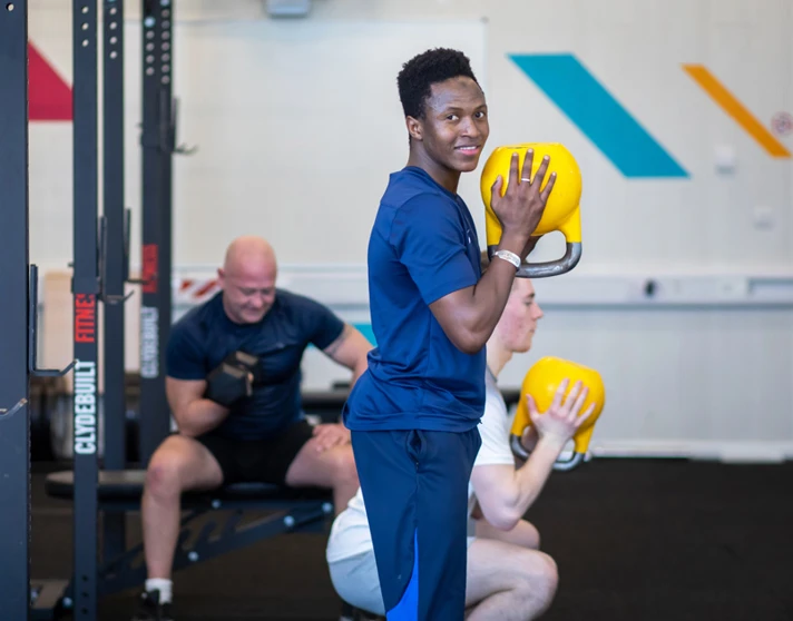 Student smiling while holding a yellow kettlebell during a strength training session at the gym. Student smiling while holding a yellow kettlebell during a strength training session at the gym.